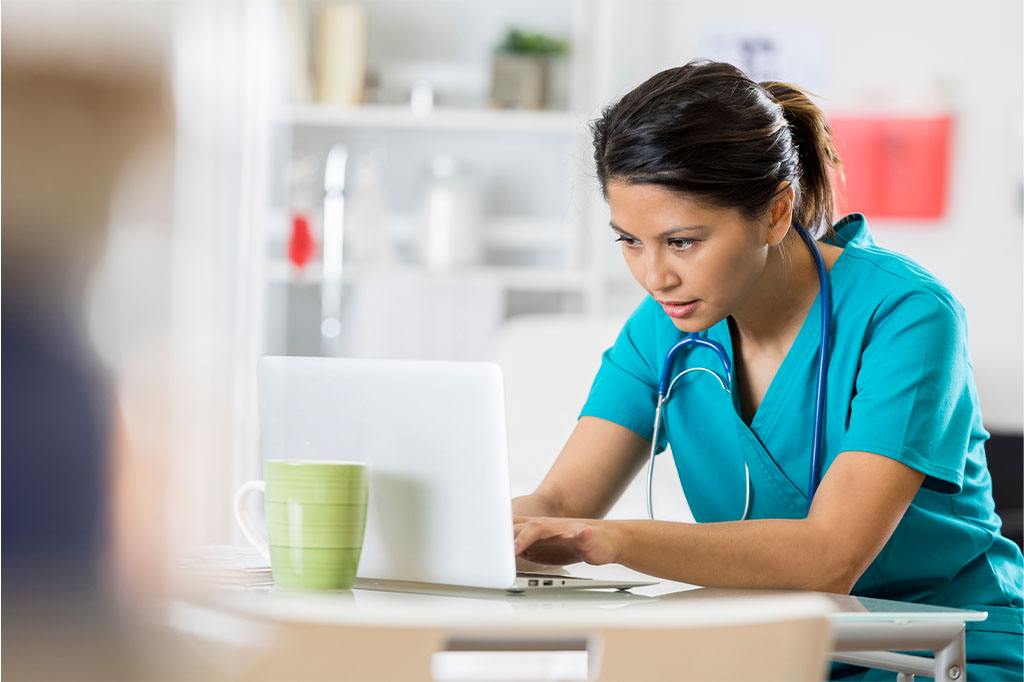 Female health care professional reads from a laptop in her office. She is wearing scrubs and a stethoscope.