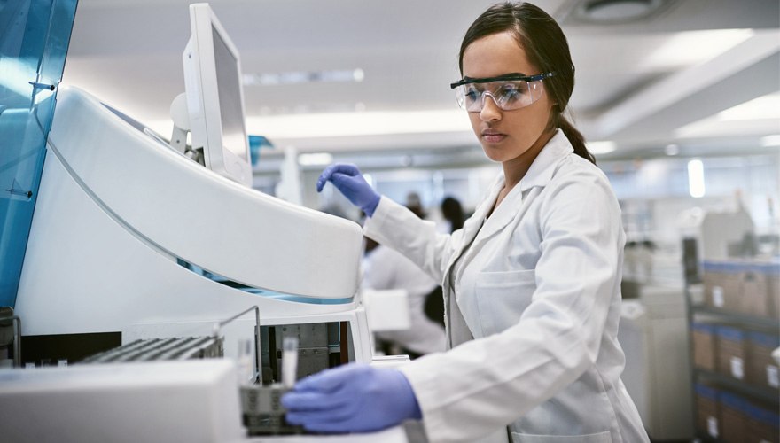 A female doctor in a white coat and googles works at a machine in a lab. 