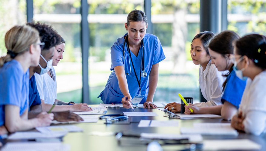 A nurse standing at the head of a table points to a document while six other health care professionals look on. 