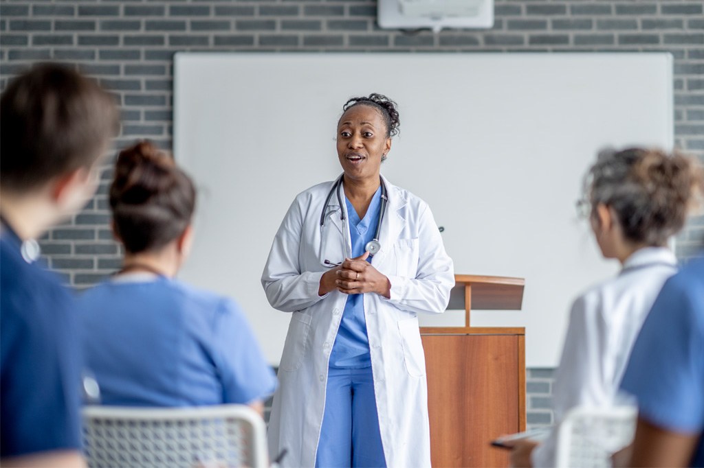 A black female doctor in a white lab coat lectures at the front of a classroom.