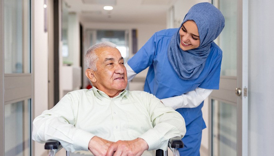 A nurse in a hijab pushes an older man in a wheelchair down a hallway. 