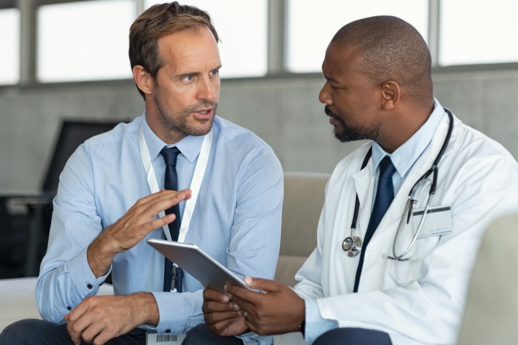 A white businessman speaks to a black doctor in a white coat. 