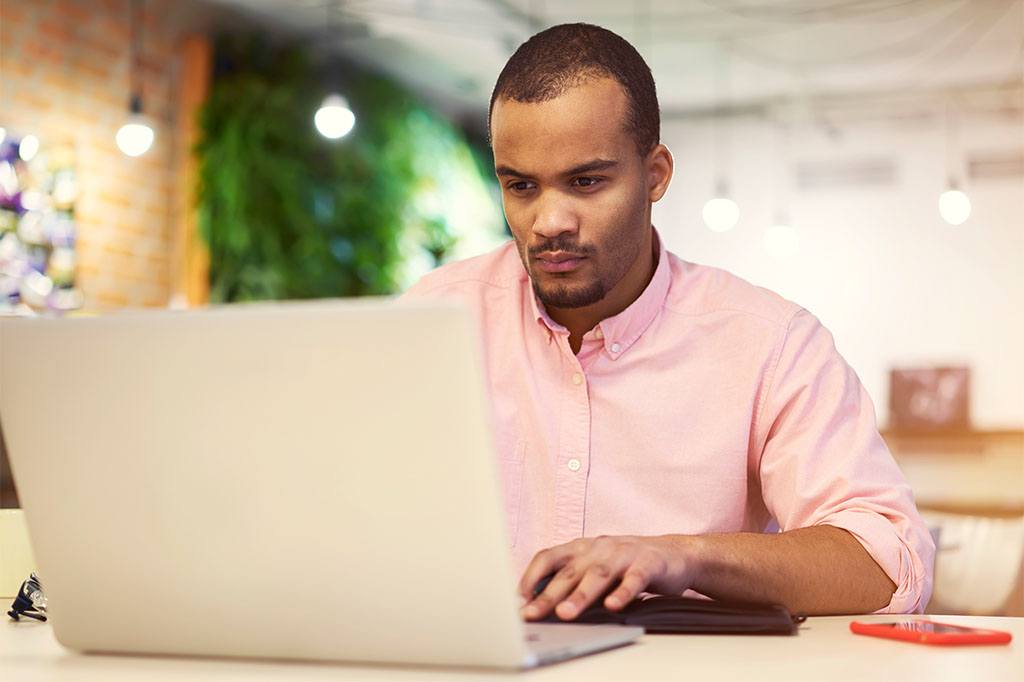 A male business professional seated at a desk reads intently from a laptop computer.